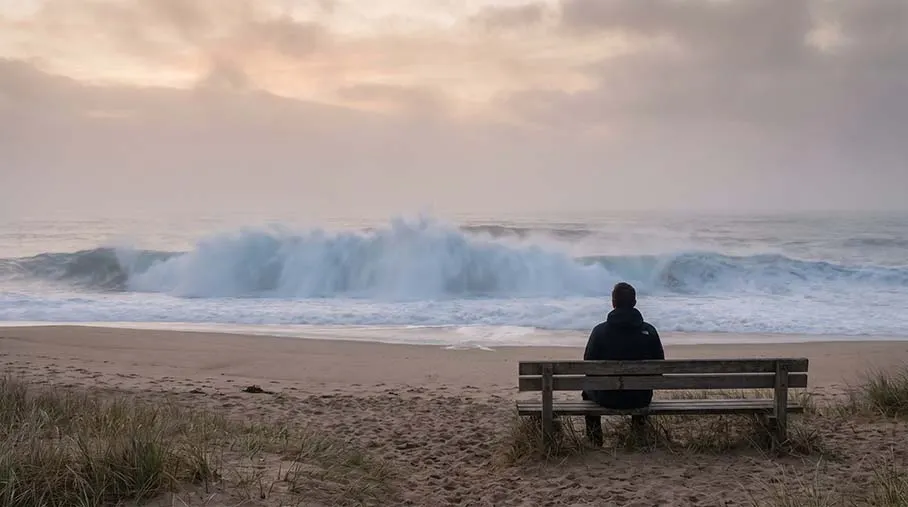 A person sitting calmly on a bench by the sea as a large wave rolls in and breaks on the shore